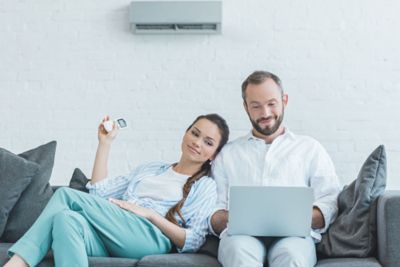 Couple sitting on a couch using a laptop while they turn on their ductless unit with a remote.