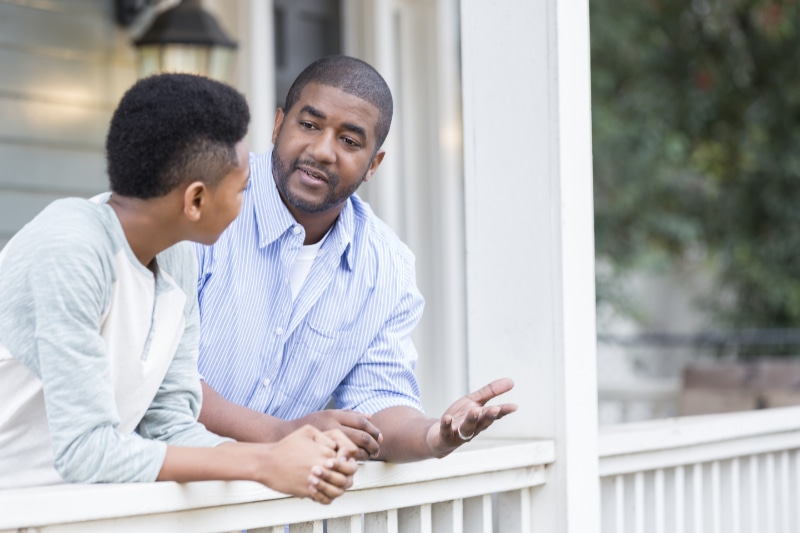 A father talks with his hands as he leans against the railing of his front porch with his preteen son and has a serious discussion.