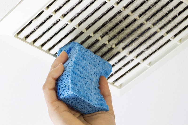 Why Air Duct Cleaning Is Important, Close up horizontal photo of female hand cleaning dirty bathroom fan vent cover with blue sponge